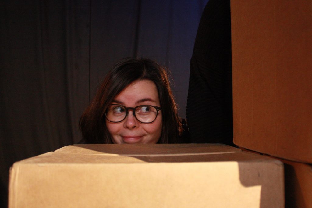 Das Gesicht einer Schauspielerin des Playbacktheater Leipzig schaut verschmitzt hinter einem Karton hervor.
