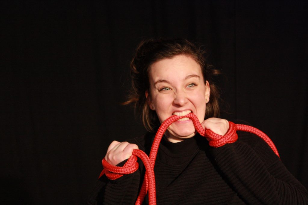 EIne junge Schauspielerin des Ensembles des Playbacktheater Leipzig ist mit einem roten Seil an den Händen gefesselt. Sie beißt auf das Seil.